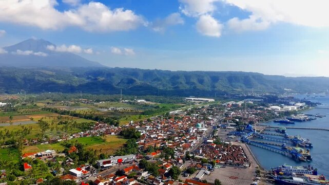 Aerial view of Ketapang Ferry Port with surrounding coastal town, lush green hills, and blue sky in Kalipuro, East Java, Indonesia.