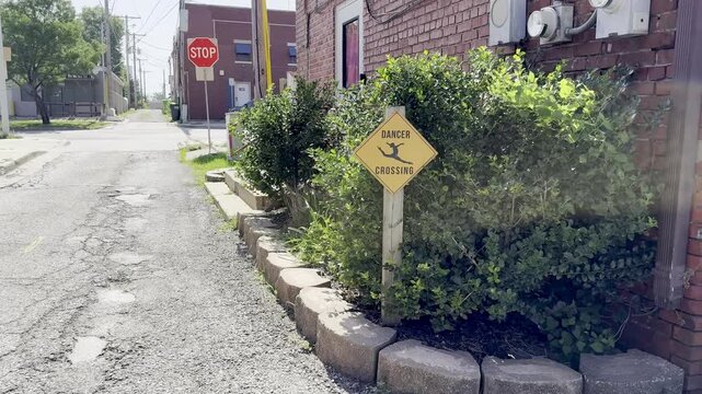 A novelty yellow diamond dancer crossing warning sign posted in a green garden bed next to a red brick building wall