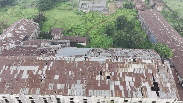 Drone footage flying right to left across the abandoned Kushtia Sugar Mill, revealing deteriorated factory buildings, damaged corrugated metal roofs, and the closed industrial complex.