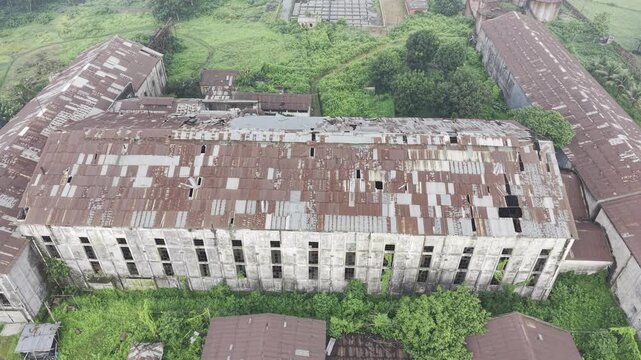 Aerial drone footage approaching the abandoned Kushtia Sugar Mill, showing deteriorated factory structures, damaged roofing, and the surrounding industrial landscape in daylight.