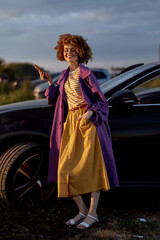 Fototapeta premium Cheerful woman outdoors, wearing a purple coat and yellow dress, smiling with confidence, sunset light, relaxed pose, soft breeze, vibrant colors, natural background