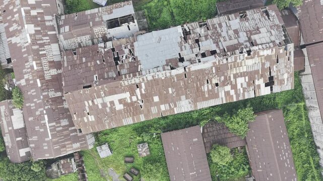 Top-down drone footage orbiting clockwise above the abandoned Kushtia Sugar Mill, revealing deteriorated factory buildings, damaged corrugated roofs, and the surrounding industrial site.