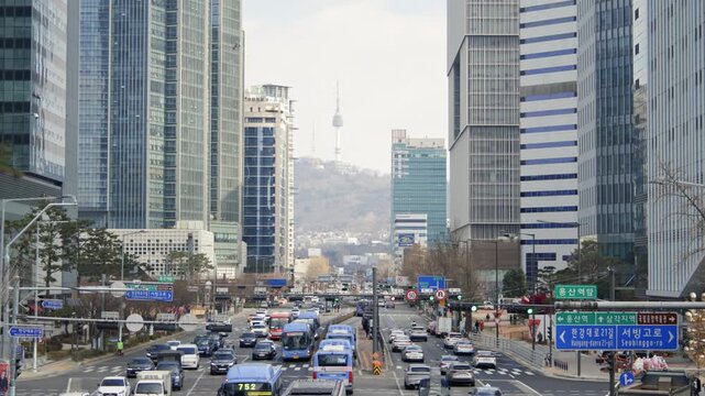 A wide view of a busy multi-lane road in downtown Seoul with modern buildings and trees lining the street on a clear day.