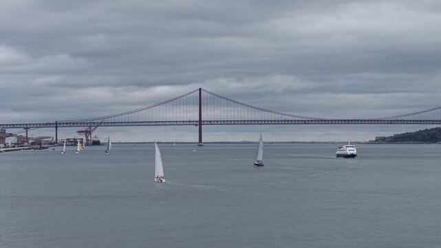 Ponte 25 de Abril suspension bridge tour by sailboats on Tagus River estuary under an overcast sky, Lisbon, Aerial