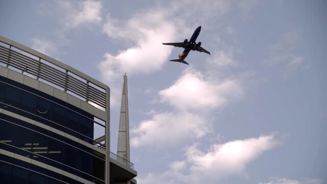 Static slow motion shot of an airplane flying over an office building. Blue sky with clouds. No people visible in this low angle wide shot.