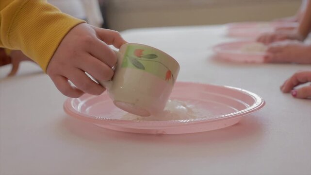 Mixing water with flour. From a cup, one hand pours a bit of water onto a small mound of flour on a pink plastic plate, where two hands are preparing to mix the two ingredients.