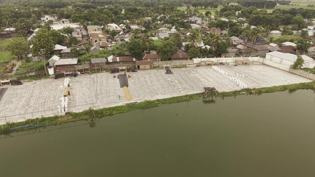 Half-orbit drone footage focusing on a rice drying yard in the Khajanagar rice processing area, showing drying surfaces, industrial buildings, ponds, and surrounding infrastructure in Bangladesh.