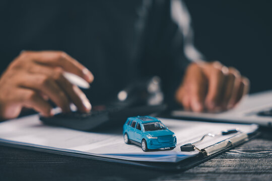 Customer signing vehicle purchase contract document for auto loan, calculating lease payment at dealership desk with blue toy car for insurance sale ownership automotive finance