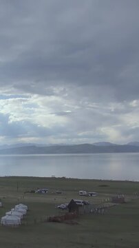 Mongolian Lake Khuvsgul glistens under a grey, cloud-filled sky. A peaceful collection of traditional gers sits along its verdant banks, reflecting the calm, ancient heart of the land.