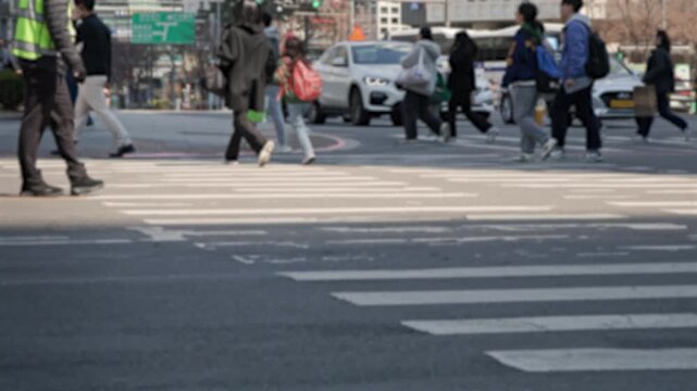A wide view of pedestrians crossing a large urban crosswalk in downtown Seoul with buildings and traffic visible in the background.