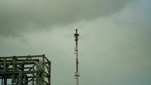 Industrial gas flare stack burning off excess gas at a petroleum refinery against a dramatic overcast sky. Concept of environmental pollution, fossil fuel energy, and chemical manufacturing.
