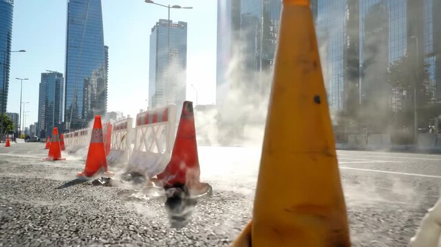 Urban construction site with orange traffic cone, white barrier, and steam rising city road, surrounded by modern glass skyscraper bright sunlight, safety warning barrier, urban street construction