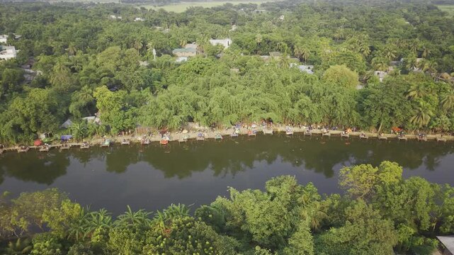 Aerial drone footage with a gentle left-to-right curve over a rural fishing competition in Bangladesh, showing people gathered along the pond bank, fishing stalls, calm water, and green countryside.