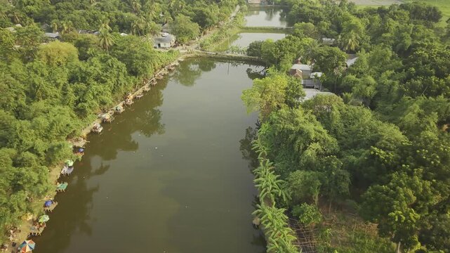 Aerial drone footage moving backward over a fishing competition at a rural pond in Bangladesh, revealing fishing stalls, people gathered by the water, calm pond surface, and lush green landscape.