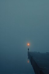 Fototapeta premium Figure stands on a dock in fog with a distant light shining in the background during early morning hours on a quiet body of water