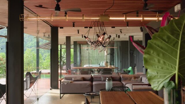 Luxurious open plan living room in tropical villa featuring wooden slat ceiling, designer pendant lights, sectional sofa, and glass walls opening to outdoor deck with lush jungle views.