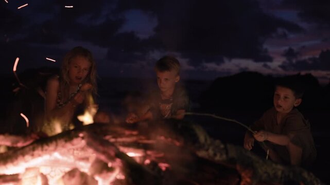 Three children gather around a glowing campfire at dusk, roasting food on sticks. Warm orange embers illuminate their faces against a dramatic twilight sky with purple and pink clouds on the horizon.
