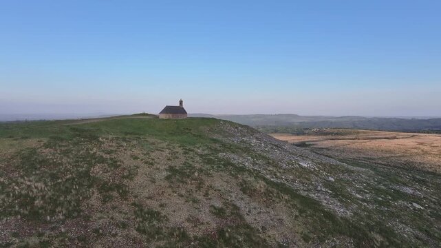 Aerial view of the historic Mont Saint-Michel de Brasparts chapel perched on a grassy hill with rocky slopes under a clear blue sky in Saint-Rivoal, Bretagne, France.