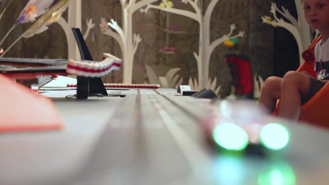 Children playing with tabletop track for cars as illuminated slot cars race along gray lanes. Young boy watches miniature vehicles with blue and green LED lights speed past decorative tree backdrop.