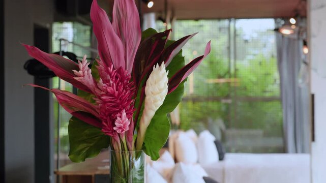 Elegant tropical flower arrangement featuring pink ginger flowers, white ginger blooms, and vibrant ti leaves in a clear glass vase. Modern interior setting with natural light and blurred background.
