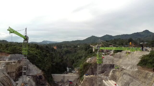 Aerial view of the PLTA Kerinci Merangin Hidro construction site featuring two large tower cranes, concrete structures, and surrounding lush forest in Batang Merangin, Jambi, Indonesia.