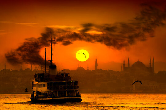 A traditional passenger ferry sailing on Bosphorus sea during dramatic sunset in Istanbul, Turkey. This scenic golden view captures historical mosque silhouettes and seagulls.
