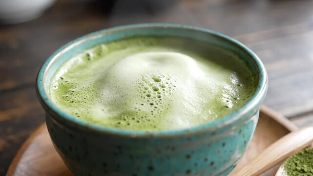 Close-up shot of frothy matcha latte in a blue bowl with matcha powder spoons and wooden plate
