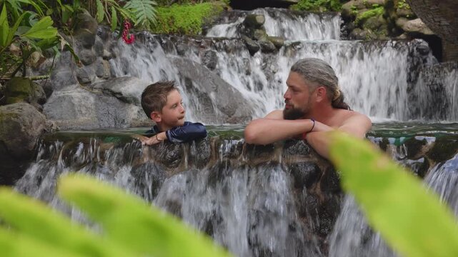 Father and son relax together in natural hot springs pool surrounded by lush tropical vegetation. Warm water cascades over rocks as they share quality time in this serene spa setting.