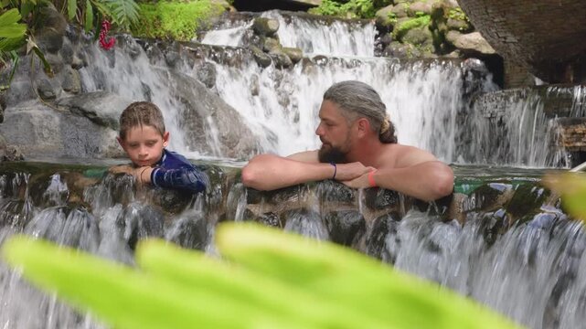 Father and son relax together in a tropical hot springs pool surrounded by lush green foliage. Water cascades over rocks as they lean on the stone edge enjoying quality time.