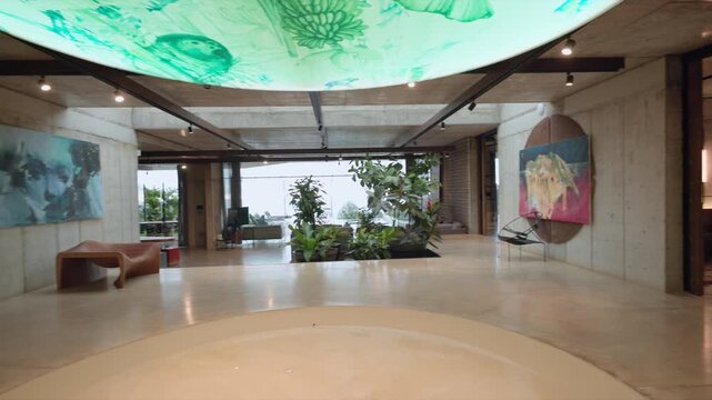 Panning shot reveals modern industrial loft space with exposed concrete walls, illuminated green artistic ceiling installation, wooden bookshelf dividers, contemporary artwork, indoor plants.
