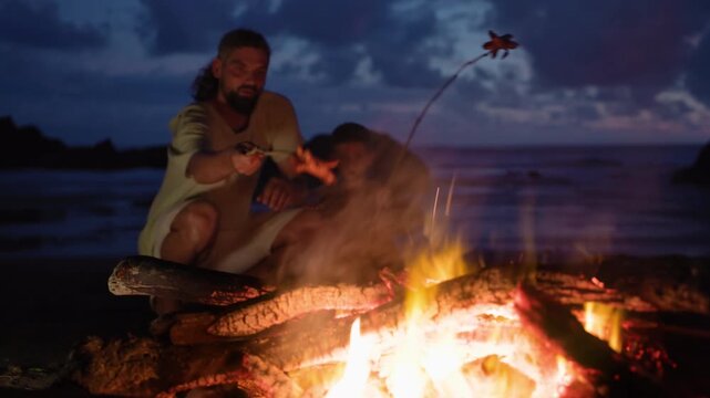 Father and son sit by campfire on ocean shore after sunset roasting sausages on sticks. Warm flames illuminate their faces against dramatic twilight sky with glowing embers and driftwood burning.