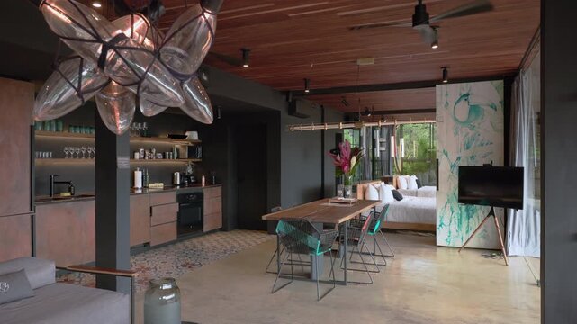 Stylish open concept loft featuring industrial design elements with wooden ceiling, modern kitchen, dining table, and cozy bedroom space with tropical plants and artistic wall decor.