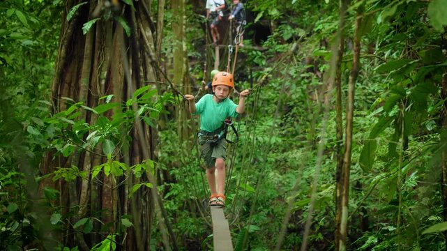 Young boy in turquoise shirt and orange helmet carefully navigates narrow wooden plank bridge while secured by zipline harness in lush tropical rainforest with dense green foliage all around.
