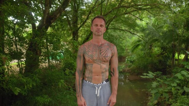 Shirtless middle-aged man with intricate geometric and tribal black ink tattoos covering his torso and arms stands calmly in a lush green tropical jungle with a peaceful river flowing behind him.