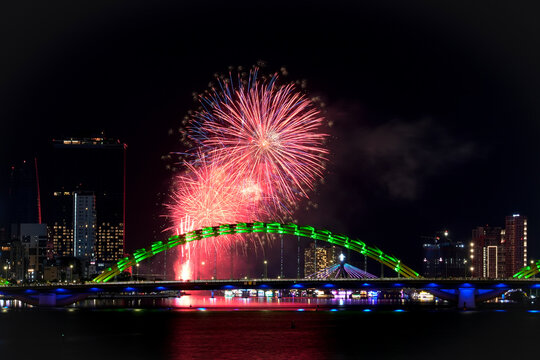 Red and pink fireworks ignite the dark night sky above Da Nang's vibrant Dragon Bridge.
