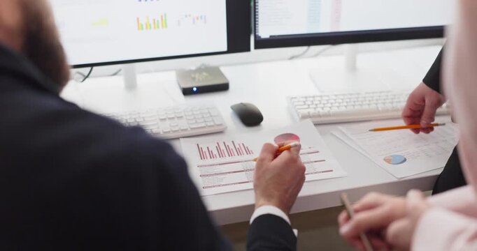Colleagues in meeting review finance charts and report. Hands with pencils highlight bar and pie metrics as monitors show data dashboards and spreadsheets. Team planning and analysis concept.
