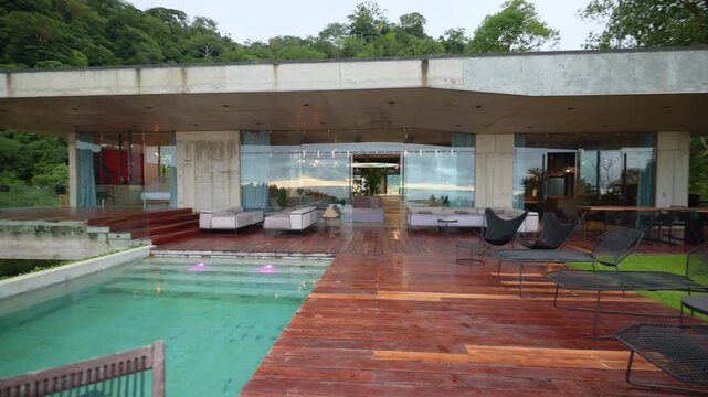 Stunning modern concrete villa featuring turquoise infinity pool and rich wooden deck terrace. Contemporary outdoor furniture and glass walls overlook lush green tropical rainforest hillside at dusk.