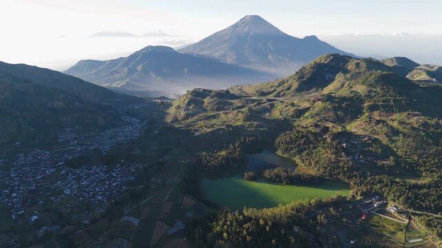Aerial view of the volcanic landscape featuring the vibrant green Telaga Warna lake surrounded by lush mountains and small village settlements in Indonesia.
