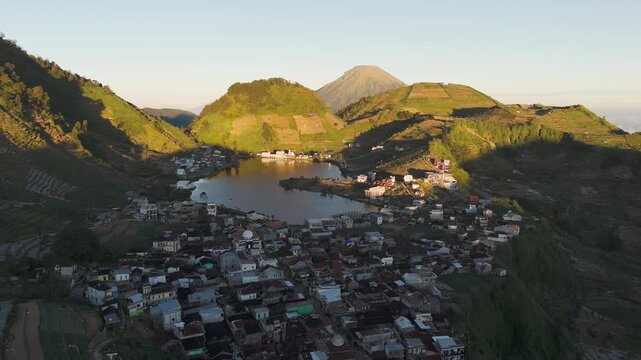 Aerial view of a village nestled by a lake surrounded by terraced hills and mountains in Indonesia.