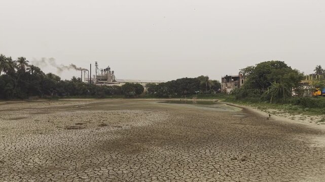Cinematic 4K drone footage rising smoothly from a cracked, dry pond to reveal the surrounding drought-stricken landscape in Bangladesh. 