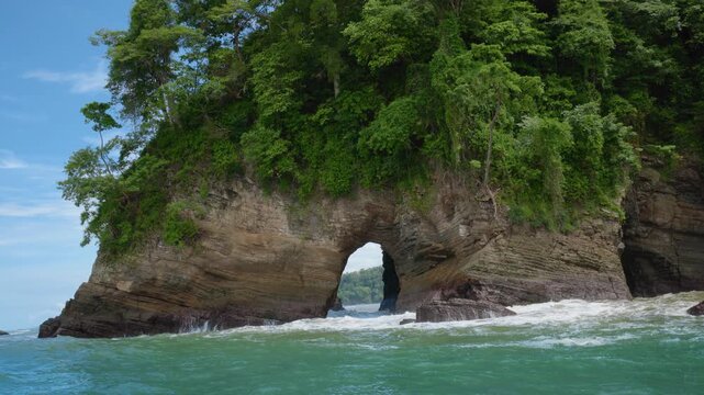 Dramatic natural rock arch formation rises from turquoise ocean waters with dense tropical rainforest covering the cliff top. Waves crash against layered sedimentary stone beneath blue sky.