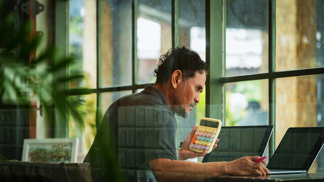 Mature businessman analyzing financial data using a calculator while reviewing reports on a laptop in a modern workspce