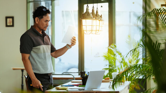 Senior businessman reviewing business documents while standing in a modern workspace. Mature entrepreneur analyzing reports