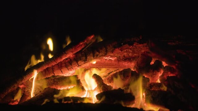 Close-up view of a campfire with glowing red embers and yellow flames dancing over burning logs. Hot coals radiate warmth as fire consumes wood against a dark night background.