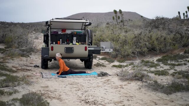 Senior woman enjoys yoga behind her teardrop camper in Baja California Mexico while camping in the desert and smiling at the moment