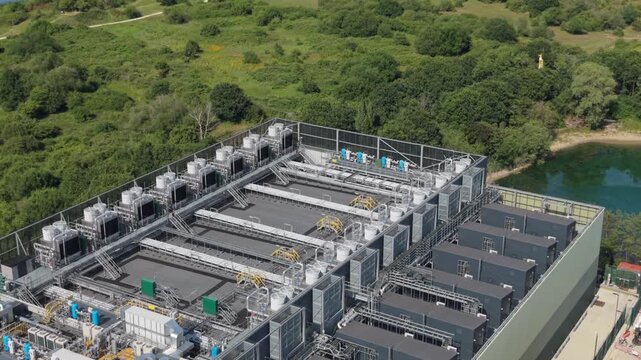 Aerial view of a large data center facility featuring rows of industrial cooling units on the roof, surrounded by lush green trees and a small pond in London, London, United Kingdom.