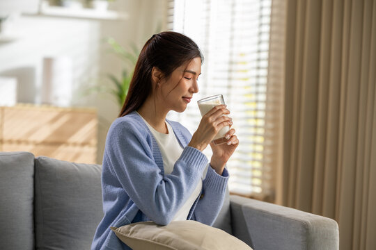 Asian woman glass drink sofa home relaxation window cardigan calm lifestyle smiling while holding beverage in bright living room with soft natural light