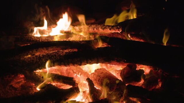 Close up view of burning campfire with glowing red embers and yellow flames dancing over charred logs. Intense heat radiates from wood coals against dark night background.