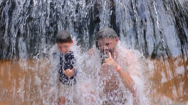 Father with son under waterfall in slow motion as cascading water splashes over happy pair. Man and boy give thumbs up gestures while enjoying refreshing summer adventure together outdoors.