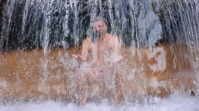 Man stands beneath a powerful natural waterfall with arms outstretched as crystal clear water cascades over his body, golden brown rock formations visible behind the rushing curtain of water.